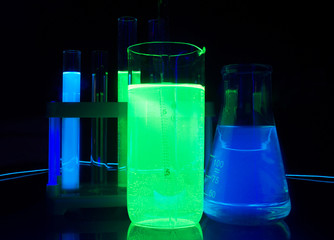 Photo composition of a laboratory bottles and test-tubes with glowing liquids standing on a glass table under ultraviolet lights.