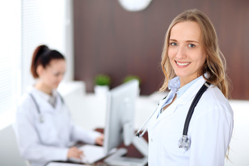 Beautiful young smiling female doctor standing in a hospital with her colleague in the background