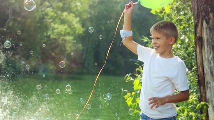 Handsome happy boy holds green balloon having fun with soap bubbles outdoors on summer in sunny day in slow motion. 1920x1080 - Powered by Adobe