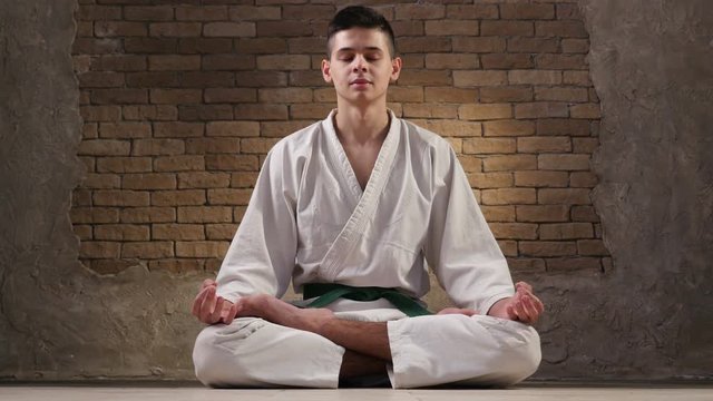 A teen boy in a white karate kimono sits in Lotus pose and meditates