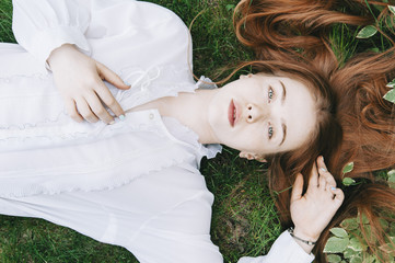 Beautiful pale faced redhead woman lying on the fresh green grass wearing a white vintage shirt,...