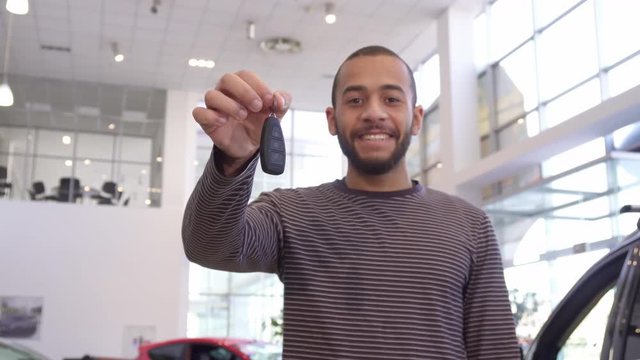 African american man showing car key at the dealership. Handsome bearded male customer smiling for the camera at the showroom. Attractive brunette guy standing near new vehicle