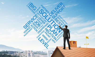Young businessman standing on house roof and writing leadership skill concept. Mixed media