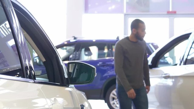 Attractive black man looking at different cars at the showroom. Handsome african american guy choosing vehicle at the dealership. Young bearded male customer putting his hand on car door
