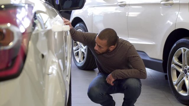 Handsome african american man looking at the car wheel at the showroom. Young bearded guy squating near the white crossover. Low shot of attractive brunette male customer putting his hand under the