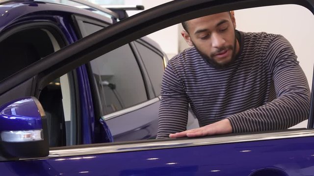 Young black man opening door of purple crossover at the showroom. Handsome african american man smiling for the camera through the car window. Attractive brunette male customer approving new vehicle