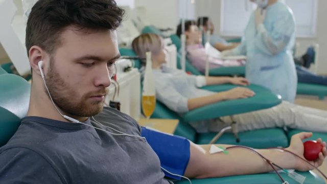 Male donor holding stress ball and listening to music on smart phone with headphones while donating blood in hospital. Nurse talking to other patients in the background