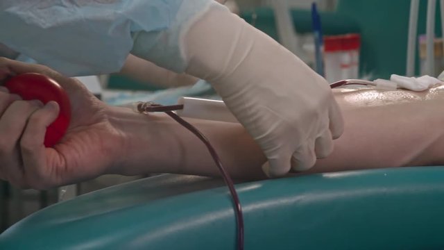 Closeup tracking shot of hands of nurse taping catheter to arm of donor gripping stress ball during procedure of collecting venous blood 