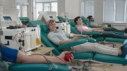 Tracking shot of young pretty woman with gauze on arm waving at camera while man holding red stress ball and using smart phone during blood donation in hospital  - Powered by Adobe