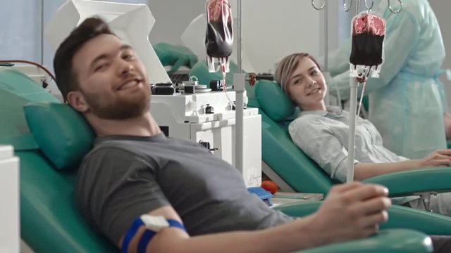 Tracking shot of young woman and man sitting in medical chairs and chatting while donating blood in hospital and then looking at camera and smiling