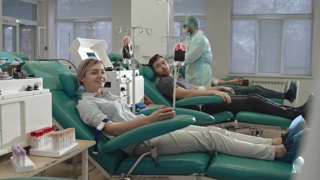 Tracking shot of young blonde woman and man sitting in medical chairs, looking at camera and smiling during blood donation process