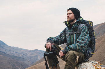 Fototapeta premium Tired bearded hipster with binoculars in his hands sits on a stone among the mountains and looks out into the distance