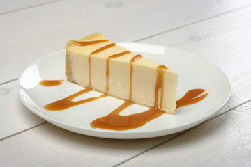 Rich piece of cheesecake, drizzled in caramel sauce on a white plate standing on wooden white table.