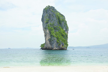 Naklejka premium Big high rock cliff filled with green vegetation surrounded by turquoise blue colored ocean water next to a tropical white sand beach with horizon view at midday, Krabi Thailand.