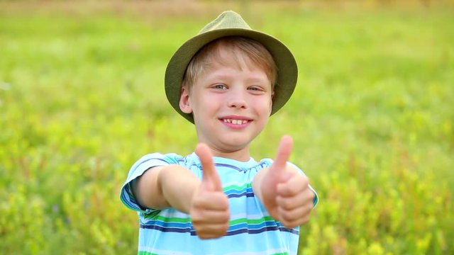 Portrait of cute funny 8 years old boy wearing green hat on summer or spring day over green grass background. Child giving two hands with thumbs up as sign of success. Real time full hd video footage.