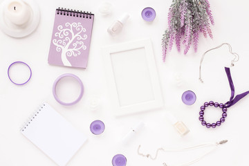 Feminine desk workspace with white frame, notebook, empty blank, stylish woman purple accessories, lilac candles, spring flowers, nail polish on white background. Flat lay, top view.