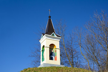 Sacral building on Lucijin Breg, Kamnica, Maribor, Slovenia. Small christian chapel on hillock. Cross as religious symbol on the top, fresco of saint person on the wall. Bare tree and clear blue sky.