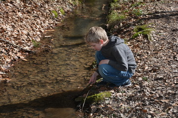 Kleiner blonder Junge an einem kleinen Bach spielt mit einem Ast im Wasser 