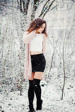 Curly brunette girl background falling snow, wear on warm knitted sweater, black mini skirt and wool stockings. Model on winter. Fashion portrait at snowy weather. Instagram toned photo.