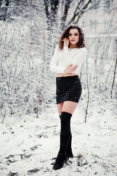 Curly brunette girl background falling snow, wear on black mini skirt and wool stockings. Model on winter. Fashion portrait at snowy weather. Instagram toned photo.