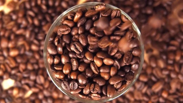 Coffee beans are poured into a glass beaker close up. Grains are poured over the edge. Slow motion.