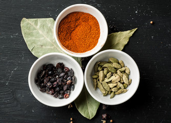 Paprika powder, barberry and cardamon seeds in white ceramic bowls on a black wooden table, top view, selective focus