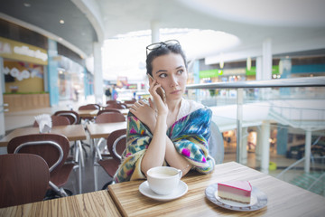 Beautiful woman is sitting in the morning at a restaurant and talking on the phone. On the wooden table in front of her are a cup of coffee and a piece of cake.