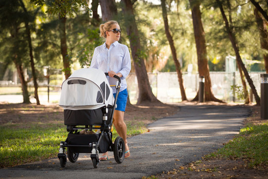 Young mother walking and pushing a stroller in the park. Mother strolling with newborn. Beautiful happy mother with pram outdoors. Summer walks in the sunny day. Girl with carriage.