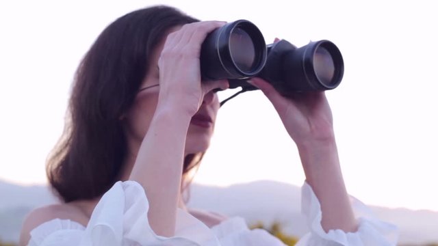 Young brunette woman watching with binoculars, looking and exploring her future. Outdoors. Shallow depth of field. 