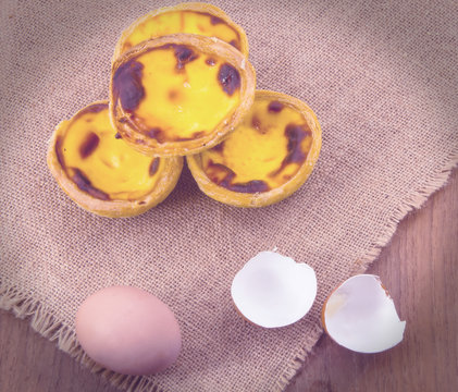 Yellow fresh egg tarts and brown chicken egg with egg shell on wooden desk with burlap cover. Retro vintage color effect
