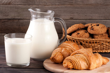 jug and glass of milk with croissants and oatmeal cookies in a wicker basket on a wooden background