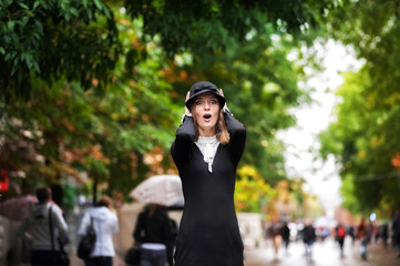 Portrait of a beautiful girl in a black dress and wearing a hat in rainy weather against the backdrop of the bright greenery of a city street. A girl with a bright umbrella is flying in the rain. Sara