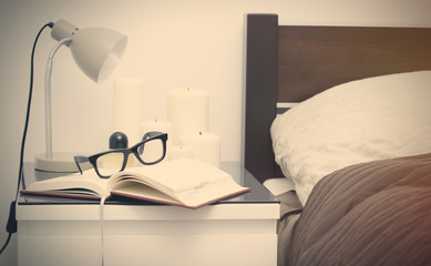 pair of glasses, lamp, candles, perfume and notebook on the table near bed in the bedroom