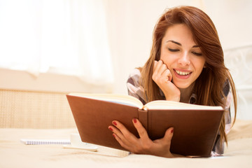 Young woman enjoying reading a book while lying on bed at home.