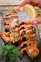 Preparing Mediterranean food. Chef hand squeezing lemon on appetizing fried shrimps served on black slate, close up view.