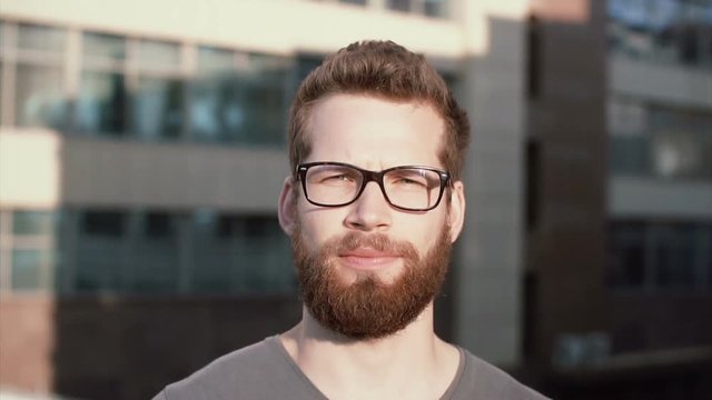 Portrait of young serious man with beard and glasses standing in front of sun, linking. Businessman stands near office.