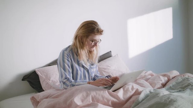 Young blonde girl in glasses wearing her pyjamas is sitting in the bed and working on her laptop from home at morning