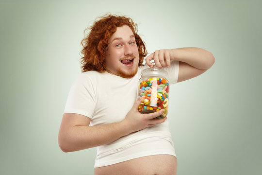 Portrait of happy excited obese young redhead male in undersized t-shirt opening jar of candies in his hands, licking lips, anticipating sweet taste of unhealthy goodies. People, obesity and gluttony