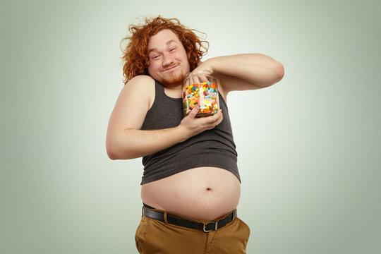 Unhealthy lifestyle, junk food, obesity and gluttony. Indoor shot of funny redhead young male, grasping handful of candies out of glass jar, having cheerful look, his big belly hanging out of pants