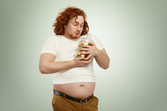 Voracious greedy fat man with red curly hair holding jar of sweets tight, looking at candies with love and affection, pouting, about to eat some. Overweight young Caucasian male posing indoors