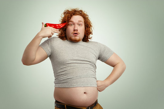 Funny overweight fat man with curly ginger hair holding big chile pepper at his temple like pistol, ready to shoot himself, having surprised expression with his belly hanging out of his grey t-shirt
