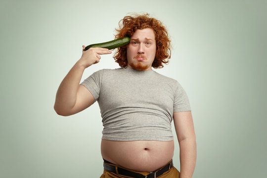 Obese overweight young redhead male ready to shoot himself out of improvised cucumber gun, having sad unhappy expression, can't stand strict vegetable diet anymore, giving up fighting against obesity