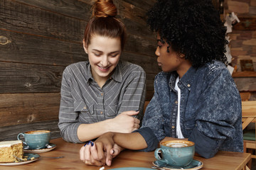 Interracial lesbian couple looking down with shy smile, holding hands during lunch at restaurant....