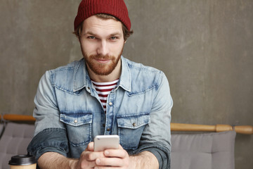 Portrait of attractive young student with thick beard wearing hat and denim jacket looking at...