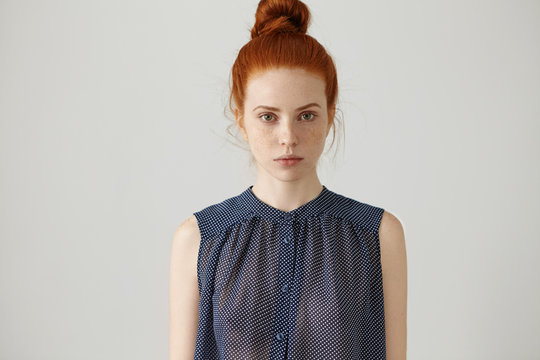 Indoor portrait of gorgeous beautiful young female model with perfect healthy freckled skin and ginger hair bun staring at camera with serious thoughtful look, dressed in spotted sleeveless shirt