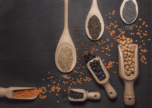 Sesame, chia and poppy seeds, red and black lentils, chickpeas and black beans with wooden spoons on black background