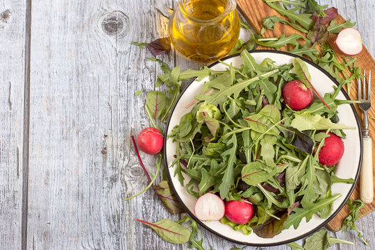
 Fresh green salad with radish. Herbs such as chard, lettuce, beet leaves on a white metal plate on a cutting board, next to a glass jug with olive oil.