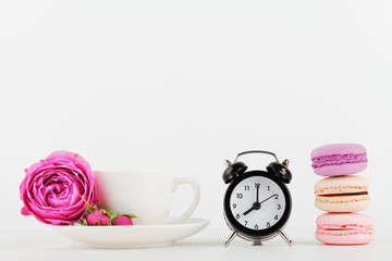 Obraz na płótnie Canvas Mockup of coffee cup with rose flower, macaroon and alarm clock on white desk with clean space for text and design your blogging.