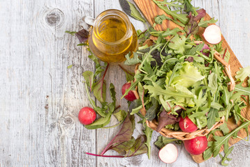  Ingredients for the preparation of spring salad. Fresh herbs such as chard, lettuce, beet leaves and a radish in a wicker basket on a cutting board, next to a glass jug with olive oil.