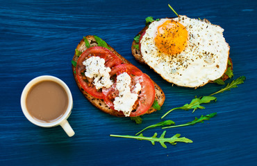 Coffee and a sandwich with feta cheese and tomatoes, fried eggs for breakfast. Dark background, top view
